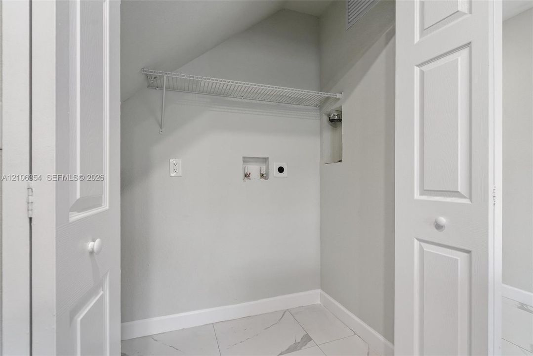 Laundry closet featuring bi-fold doors, white large-format stone-finish tile flooring, a wall-mounted wire storage shelf, and dedicated water and electrical hookups