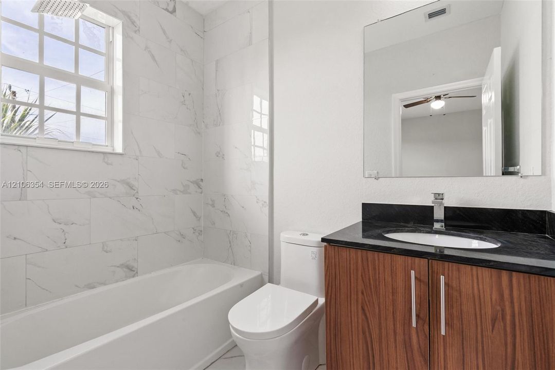 Bathroom featuring large-format veined tile tub surround, dark countertop with an integrated basin, wood-finish cabinetry, and a wall-mounted mirror