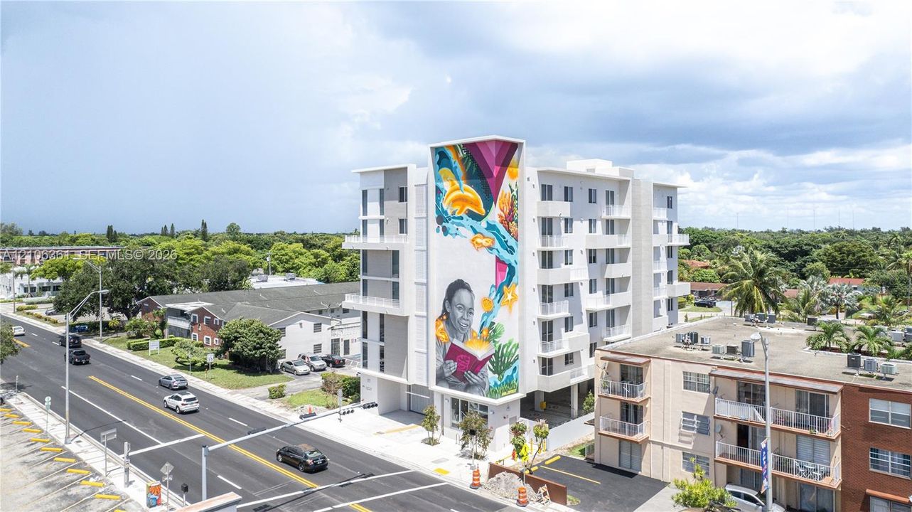 Multi-story residential building featuring a large-scale exterior mural, private balconies, and ground-level covered parking