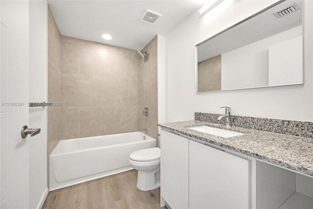 Bathroom featuring a white bathtub, beige tile tub surround, wood-finish flooring, stone-surface vanity countertop with a rectangular undermount sink, and a full-width wall-mounted mirror