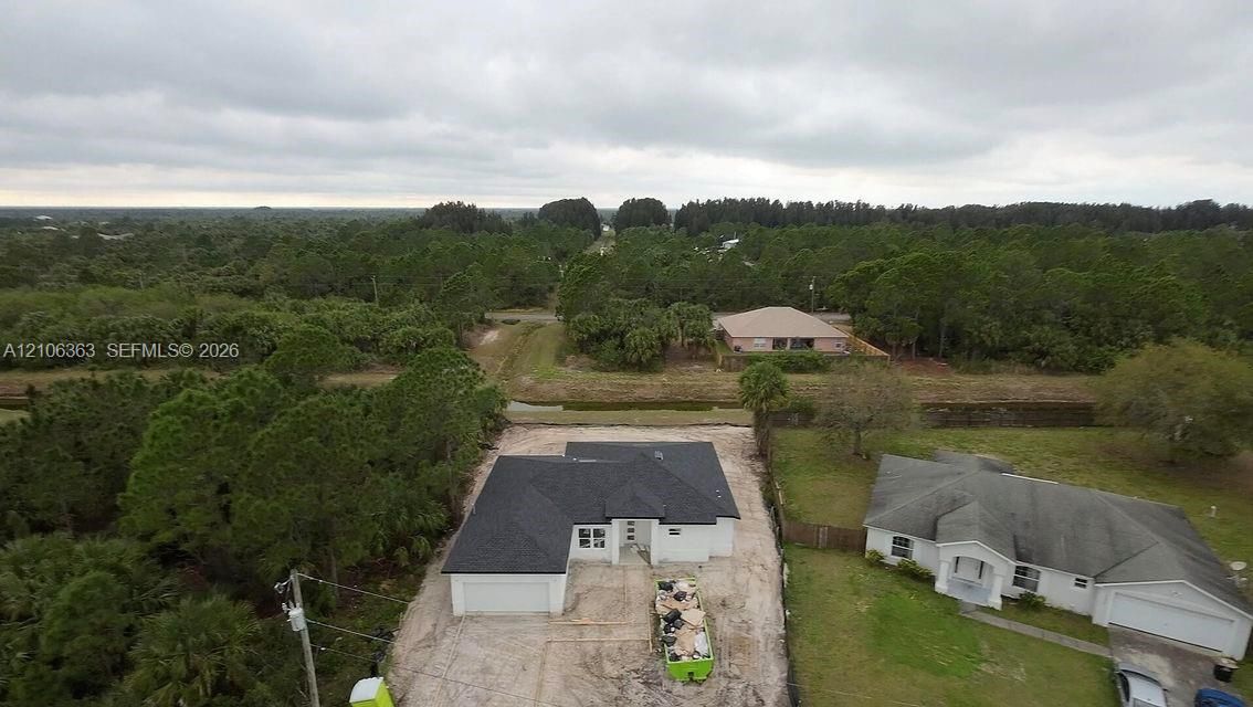Aerial perspective of a single-story residence featuring a dark shingle roof, exterior white siding, and a multi-car garage, positioned on a lot with expansive surrounding woodland