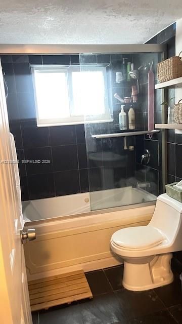 Bathtub and shower combination featuring dark square tile surround, sliding glass door, white porcelain toilet, and wall-mounted shelving
