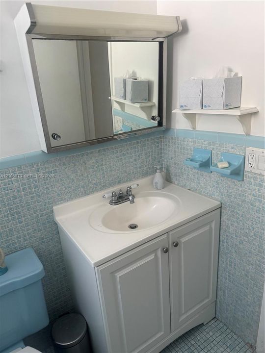 Vanity cabinet with a white basin and chrome-finish dual-handle faucet, metal-frame medicine cabinet, and wall-mounted shelving featuring blue mosaic tile detailing