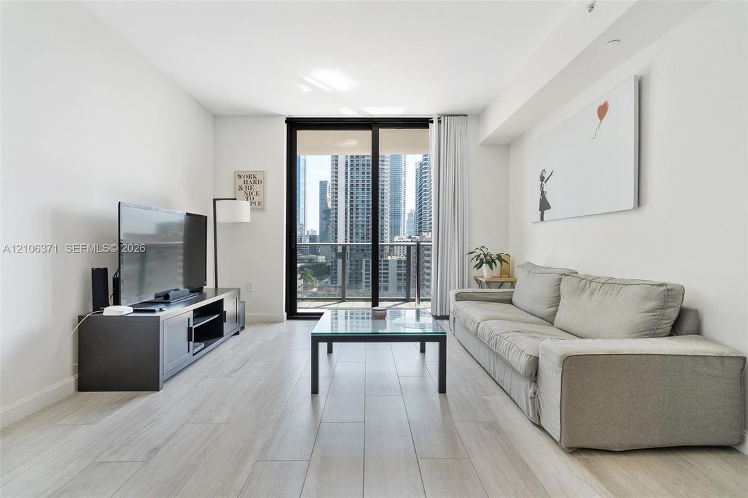Living area featuring light-toned wood-finish flooring, floor-to-ceiling sliding glass doors, and a private balcony with metallic railing