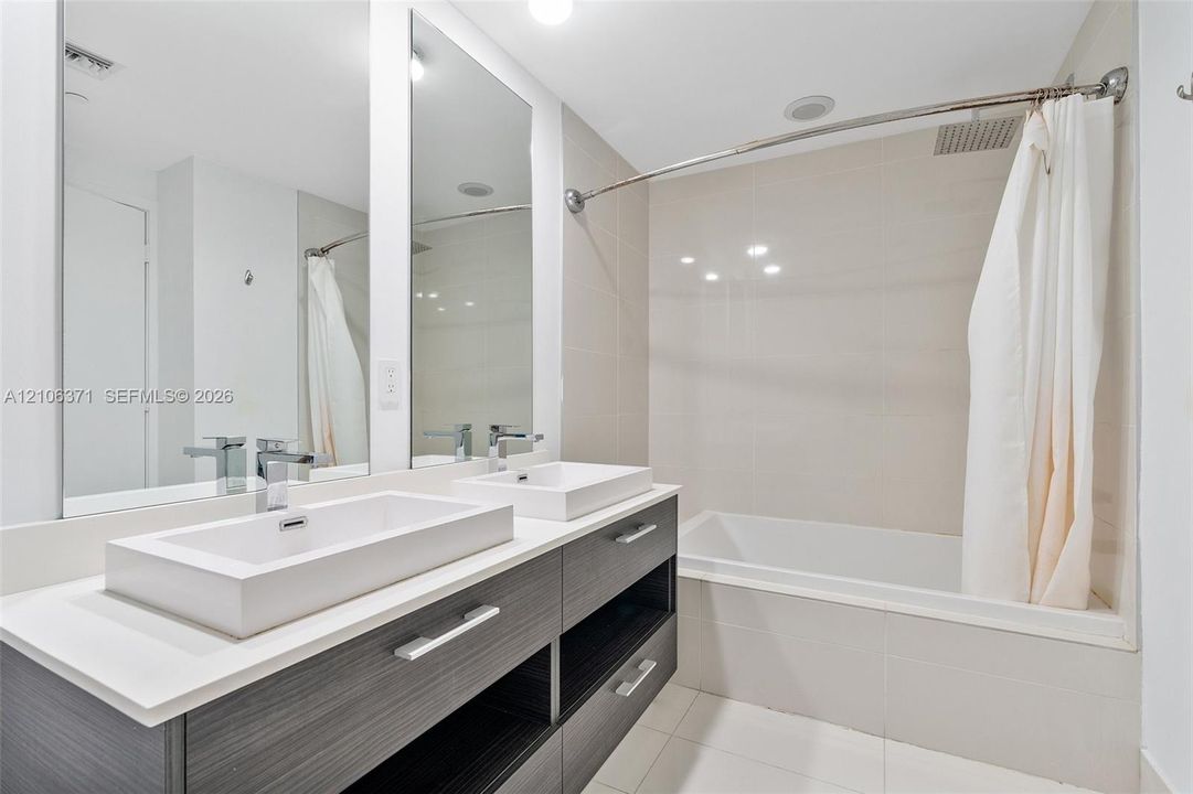 Double vanity featuring a light-toned countertop with rectangular vessel sinks, dark-finish cabinetry, full-width mirror, and a tile-surround bathtub