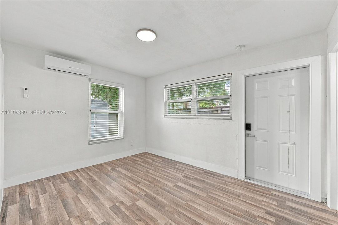 Wood-finish flooring, white walls, dual windows with horizontal blinds, wall-mounted ductless air conditioning unit, and white six-panel exterior door