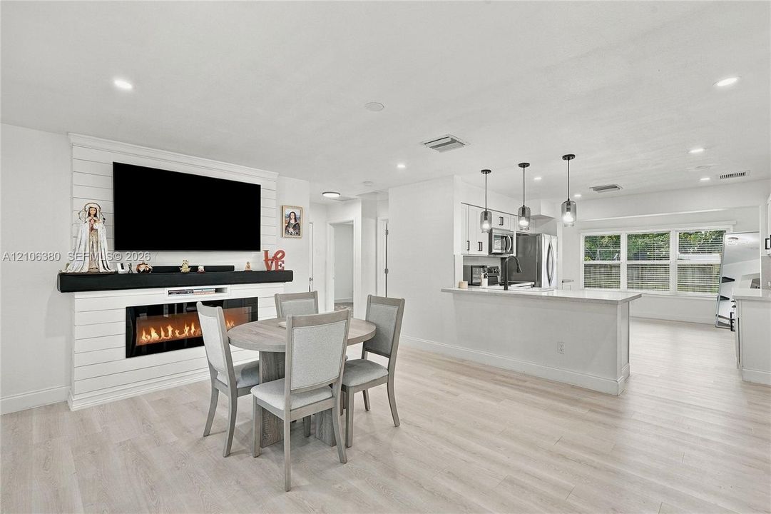 Open-concept interior featuring shiplap fireplace surround, electric fireplace insert, wood-finish flooring, kitchen island with pendant lighting, and multi-pane windows