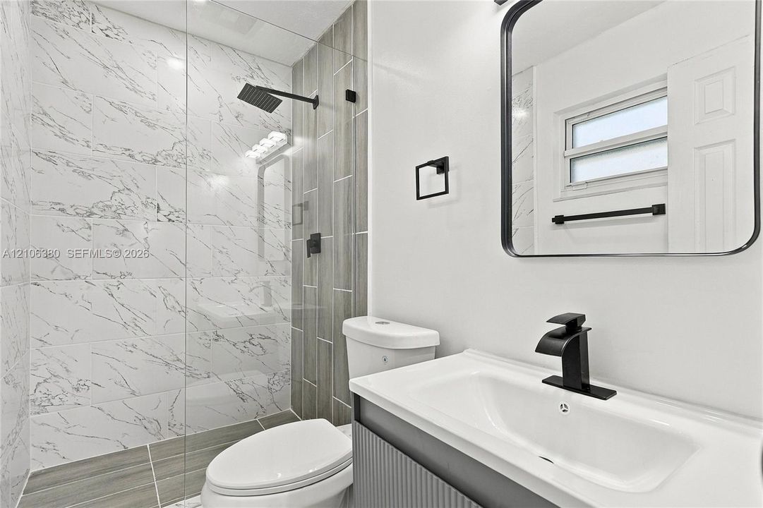 Bathroom featuring a glass-enclosed shower with marbled tile, a rain-style showerhead, wood-finish wall tile accents, a vanity with a rectangular basin and matte black faucet, and a black-framed mirror