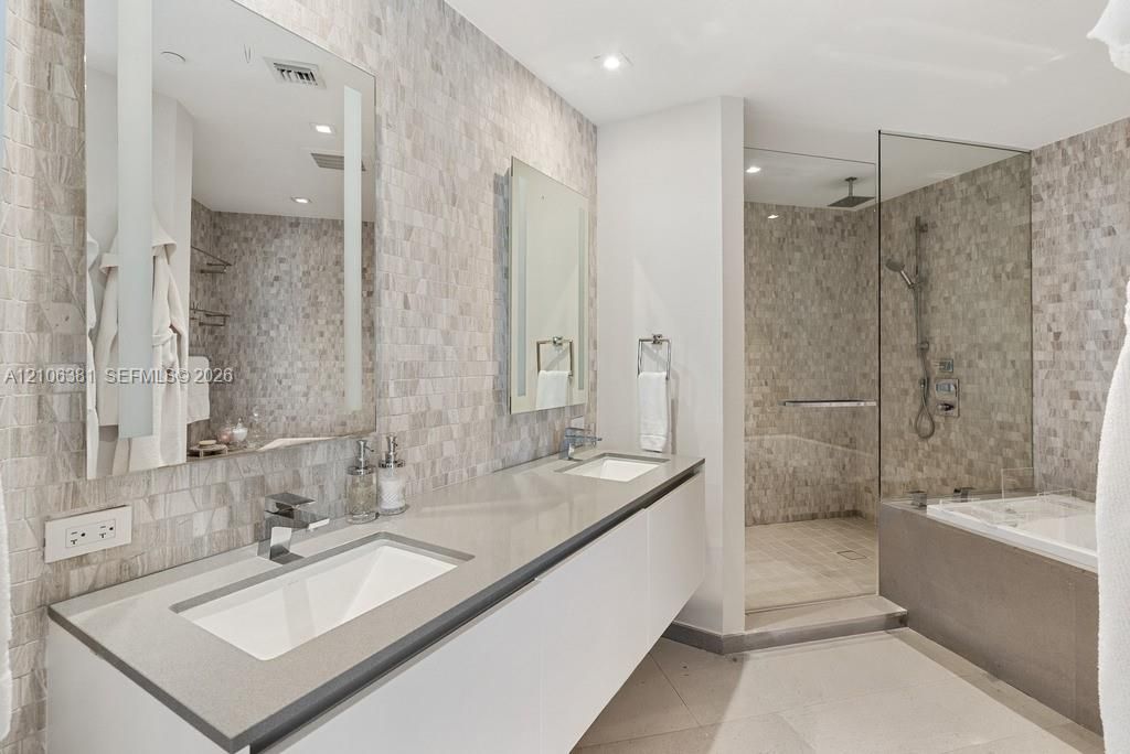 Floating dual-sink vanity with a solid surface countertop, textured stone tile wall accents, glass-enclosed walk-in shower, and a built-in bathtub