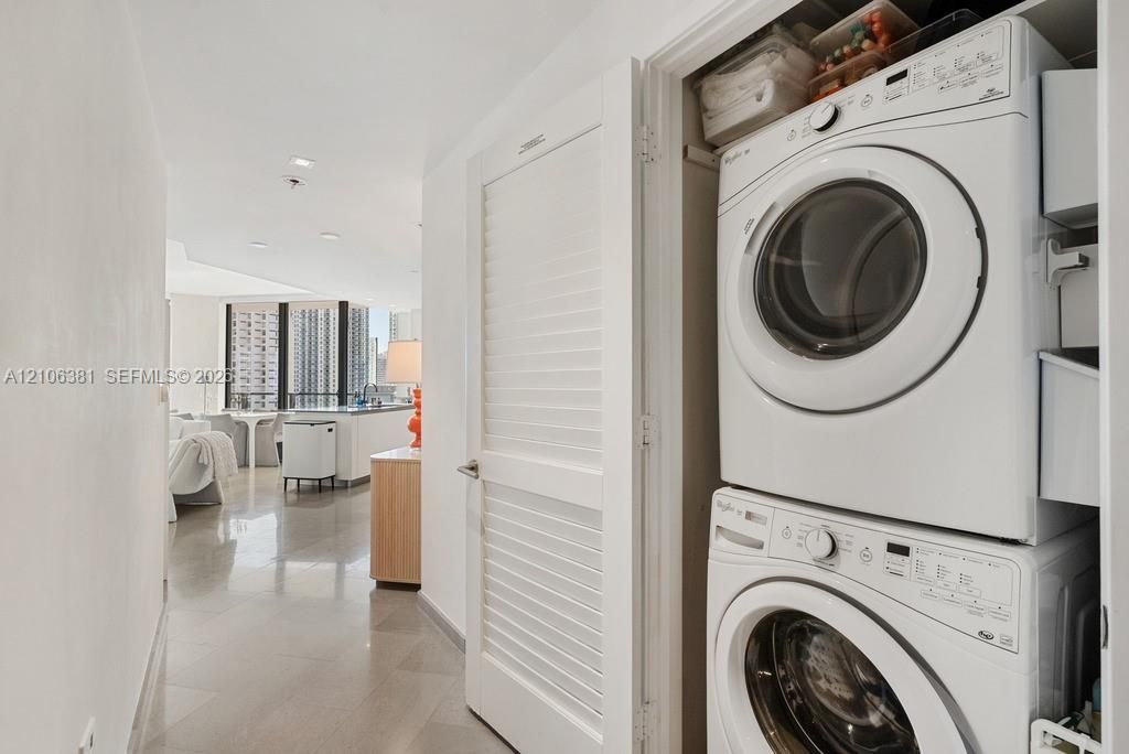 Stackable washer and dryer unit enclosed by white louvered doors, featuring tile flooring and recessed ceiling lighting