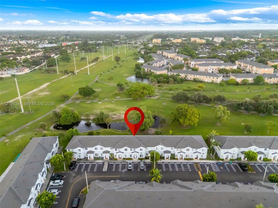 Aerial perspective featuring multi-story residential buildings, paved surface parking, a pond, and expansive green space