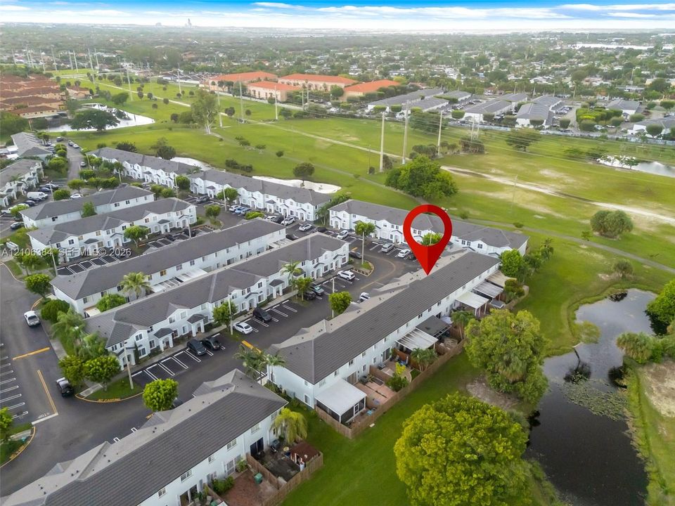 Aerial perspective of a multi-unit residential property featuring dark-toned shingle roofing, paved surface parking, and exterior rear patios, situated on a grass-covered landscape with a natural water feature