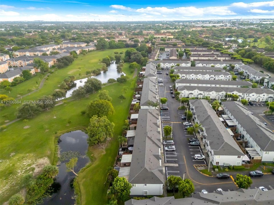 Aerial perspective of a multi-unit residential community featuring pitched shingle roofs, paved asphalt parking areas, expansive common green space, and natural water features