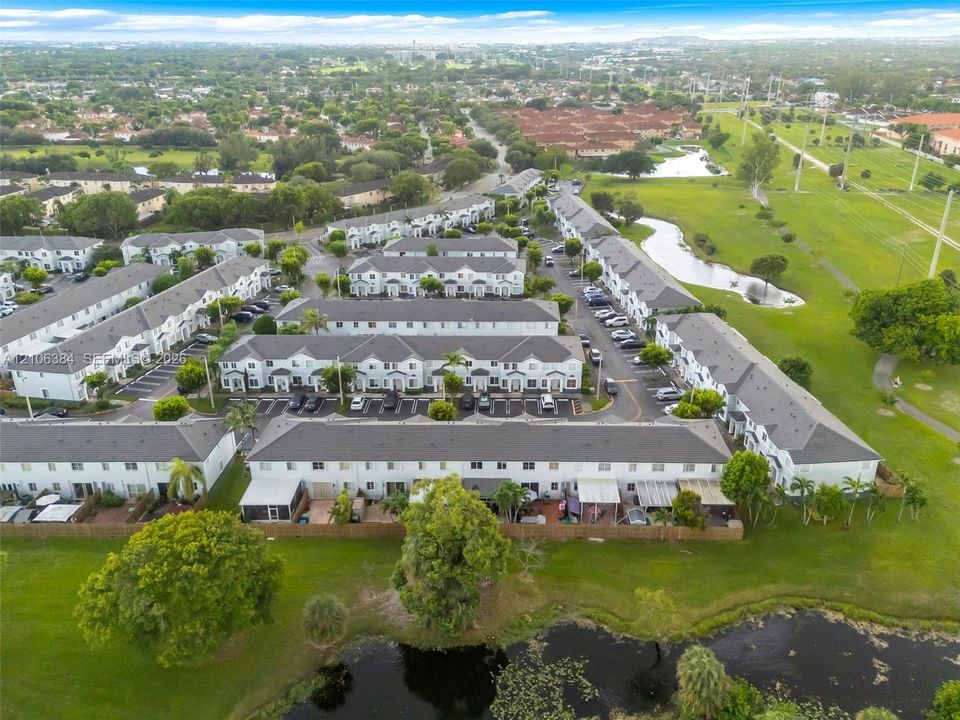 Aerial perspective of multi-family residences featuring grey shingle roofing, exterior light-toned finishes, dedicated parking spaces, and expansive green space with water features