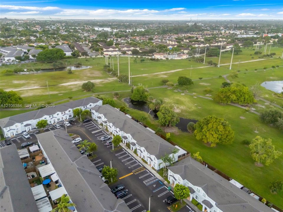 Aerial perspective of a multi-unit residential development featuring a gray shingle roofline, paved parking area with designated stalls, and extensive green space with established trees and water features
