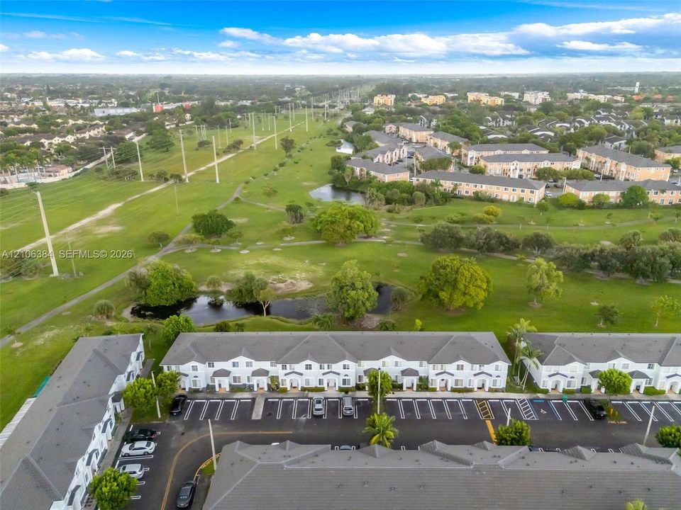 Aerial perspective of multi-family structures featuring peaked grey-tone roofs and white siding, including dedicated asphalt parking areas and proximity to expansive green space with mature trees and a retention pond