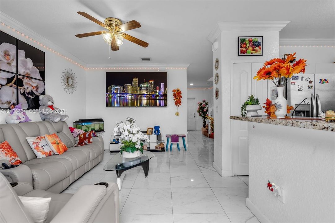 Open-concept interior featuring light-toned tile flooring, crown molding, a multi-blade ceiling fan with integrated lighting, and a breakfast bar topped with stone-finish surfacing