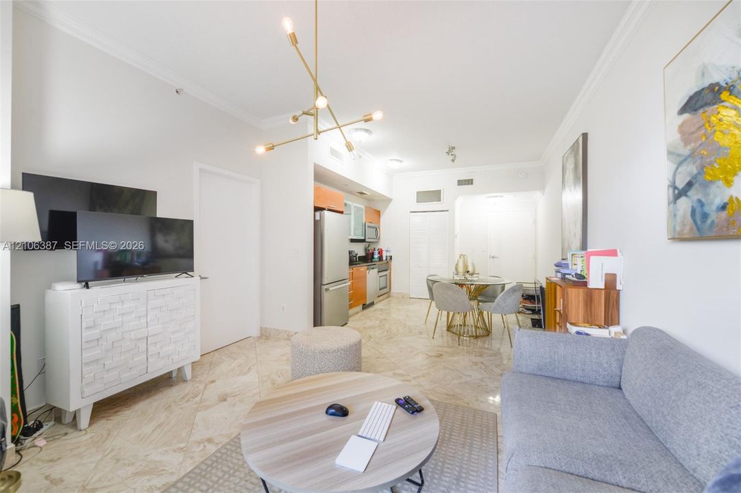 Open-concept interior featuring large-format tile flooring, a modern multi-arm brass chandelier, crown molding, and a kitchen equipped with stainless steel appliances