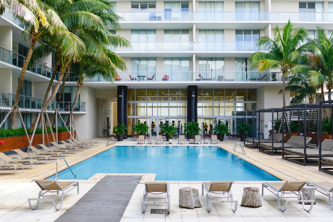 Swimming pool featuring a paved deck, perimeter palm trees, tiered building levels with private balconies, and glass-walled common area