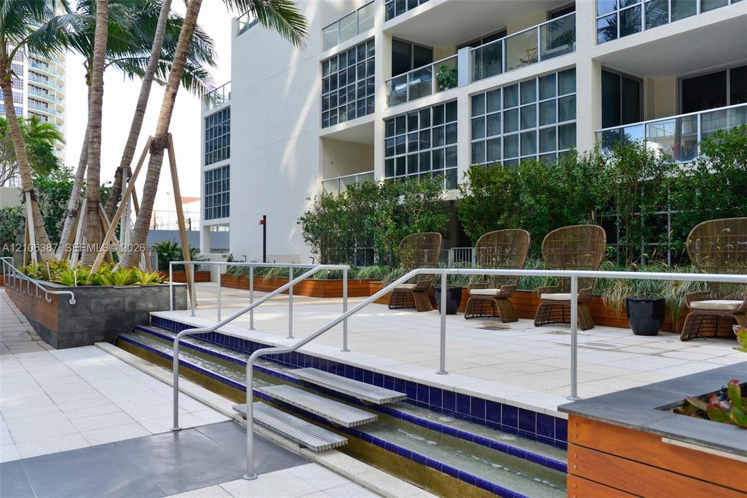 Outdoor terrace featuring tiered tile decking, a linear water feature with blue tile accents, multi-level metal handrails, and integrated planter boxes with wood-finish siding