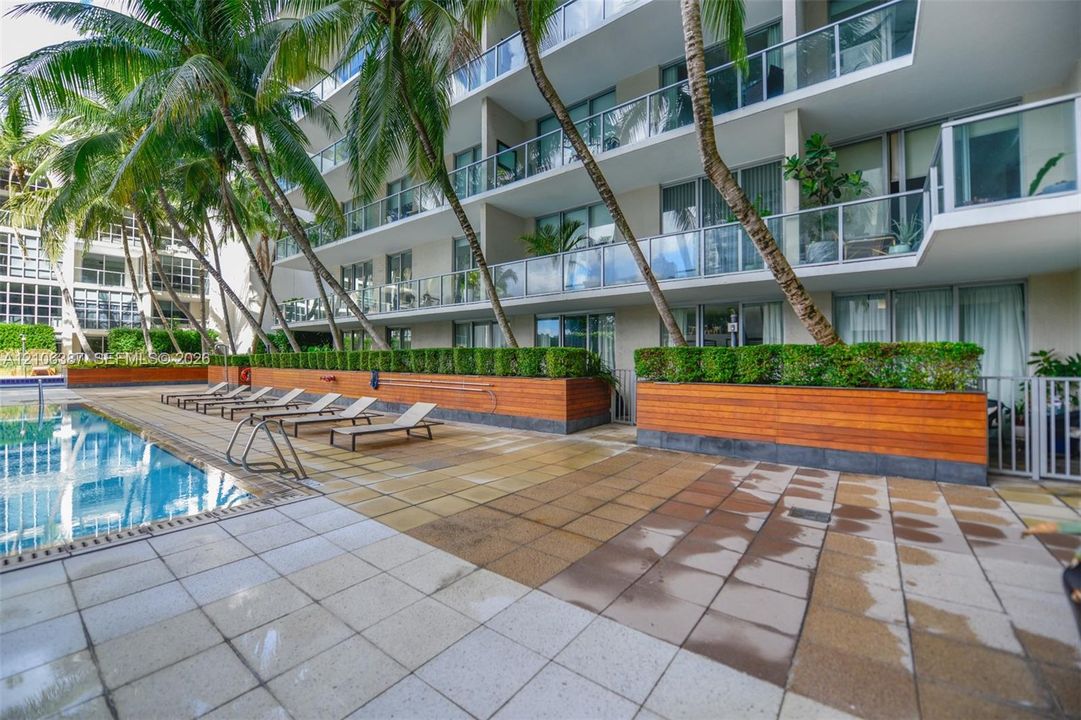 Multi-story residential building featuring private balconies with glass railings, an outdoor swimming pool, stone-tiled patio, and wood-paneled planter boxes