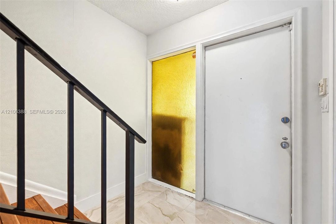 Entryway featuring a solid white door, a floor-to-ceiling amber-tinted sidelight, stone-finish tile flooring, and a metal staircase railing