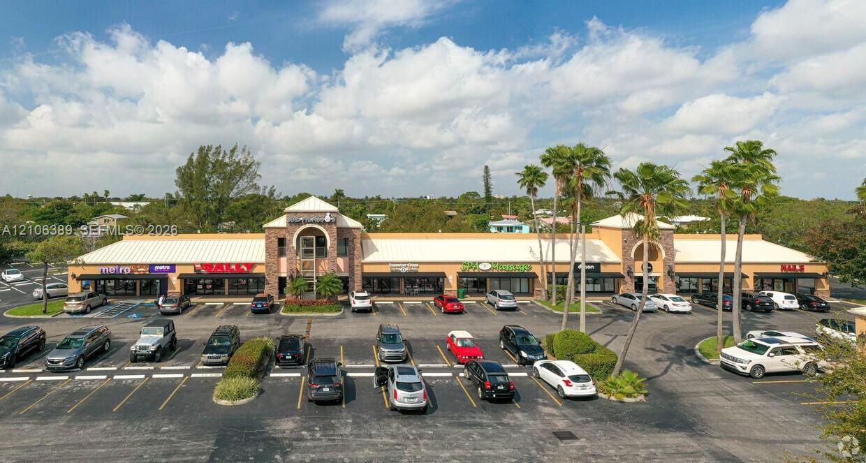 Commercial strip center featuring a masonry exterior, prominent arched entryway, metal roofing, expansive asphalt parking area, and mature palm landscaping