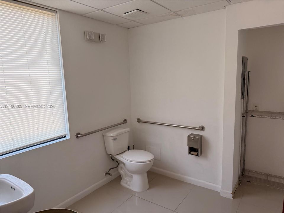 Restroom featuring large-format tile flooring, a porcelain toilet, stainless steel wall-mounted grab bars, a built-in metal paper dispenser, and a window with horizontal blinds