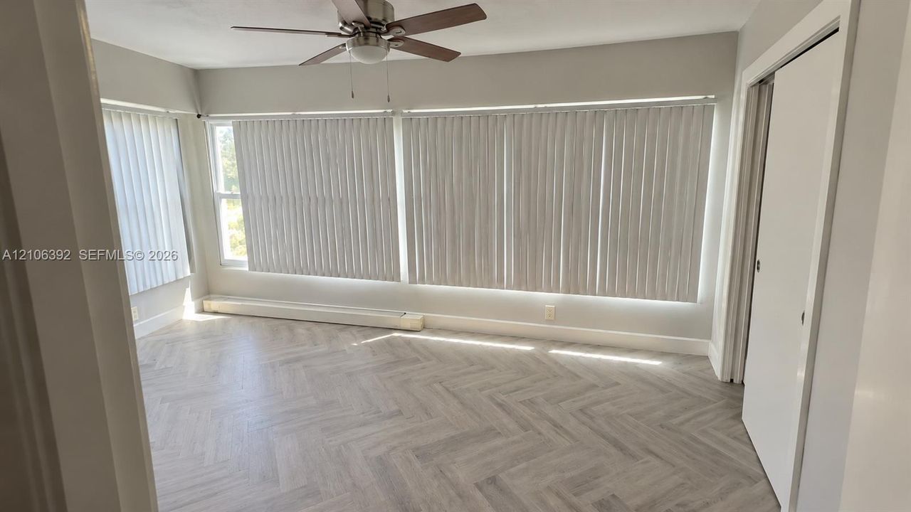 Herringbone-pattern wood-finish flooring, extensive floor-to-ceiling windows with vertical blinds, and a neutral-toned ceiling fan with integrated lighting