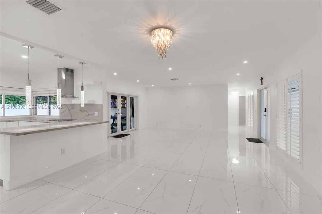 Open interior space featuring polished tile flooring, recessed lighting, a multi-tier crystal flush-mount light fixture, white plantation shutters, and a breakfast bar with stone-surfaced countertops