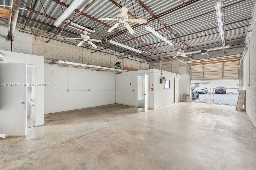 Open-concept utility space featuring high corrugated metal ceilings, polished concrete flooring, industrial overhead lighting, multiple ceiling fans, and a roll-up bay door