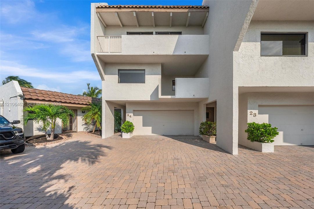 Multi-level stucco exterior featuring a paver stone driveway, attached garage, recessed balconies, and exposed beam ceiling details
