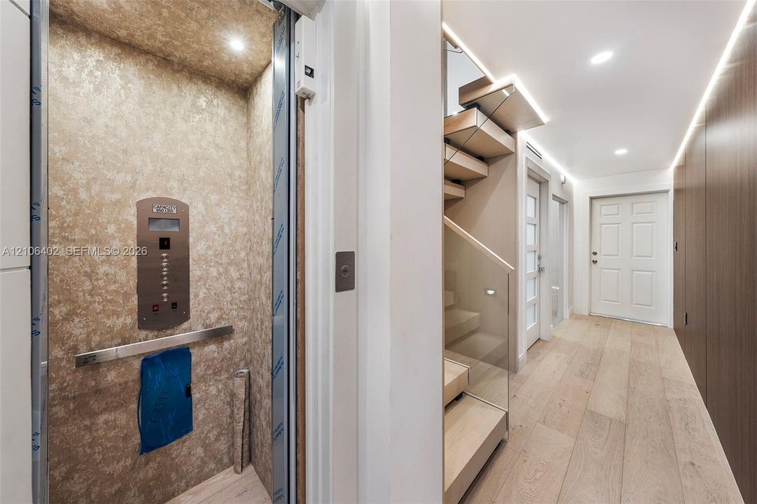Private elevator featuring metallic-finish wall paneling and a stainless steel control panel, wood-finish flooring, staircase with glass railings and wood treads, recessed lighting, and wall-mounted linear lighting