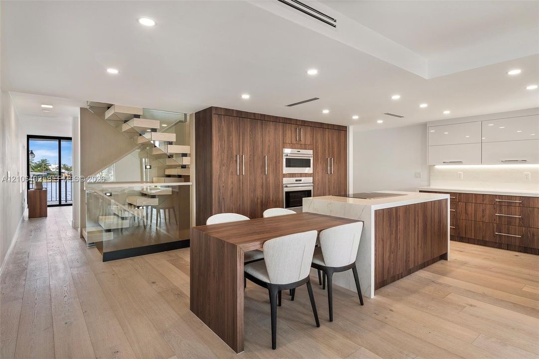 Modern kitchen featuring wood-veneer cabinetry, stone-topped center island with integrated cooktop, recessed lighting, wood-finish flooring, and a floating staircase with glass paneling