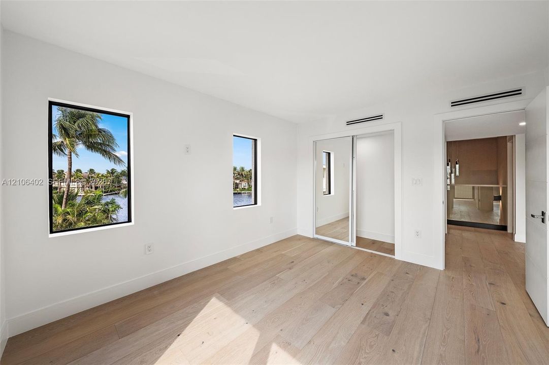 Interior room featuring wide-plank wood-finish flooring, floor-to-ceiling mirror-panel sliding closet doors, and multiple windows with water and palm tree scenery