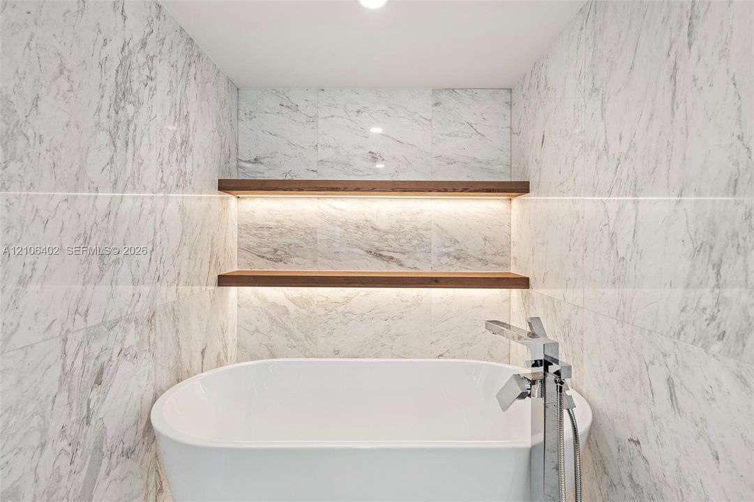 Freestanding soaking tub featuring a chrome floor-mounted faucet, full-height stone-finish wall tile, and built-in wood-tone shelving with recessed accent lighting