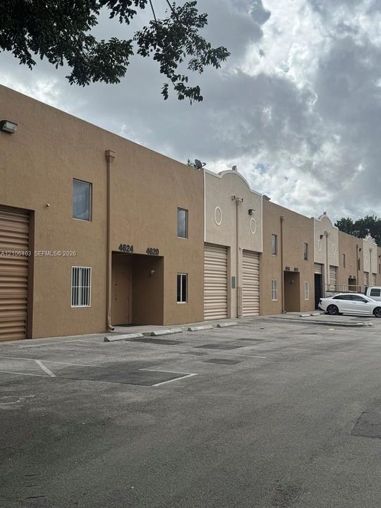 Stucco exterior commercial complex featuring roll-up bay doors, individual unit entries, concrete paved parking, and wall-mounted exterior lighting