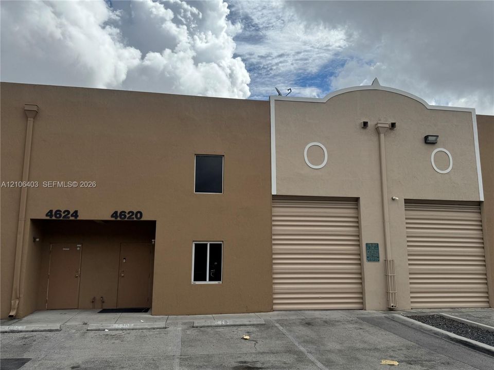 Stucco facade featuring two roll-up metal bay doors, dual entry doors, exterior wall-mounted lighting, and a decorative arched parapet