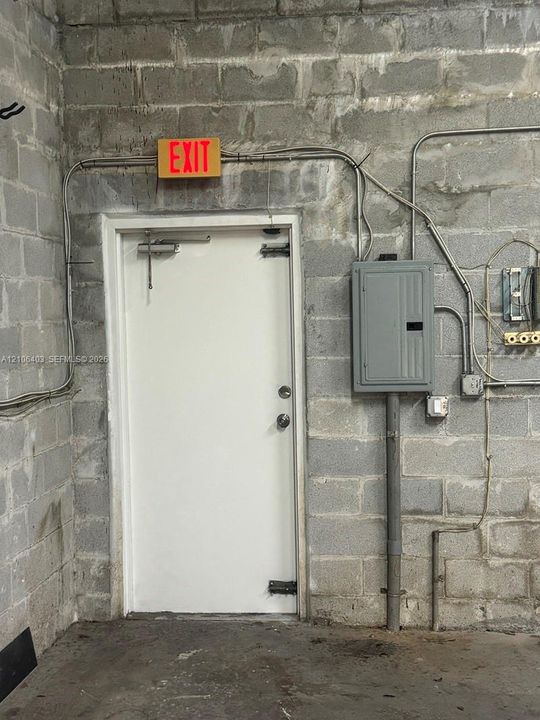 Concrete block wall construction featuring a steel entry door with overhead hydraulic closer, surface-mounted electrical service panel, and illuminated exit sign