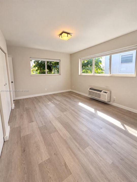 Interior space featuring light-toned wood-finish flooring, dual windows with exterior greenery, and an integrated wall-mounted climate control unit