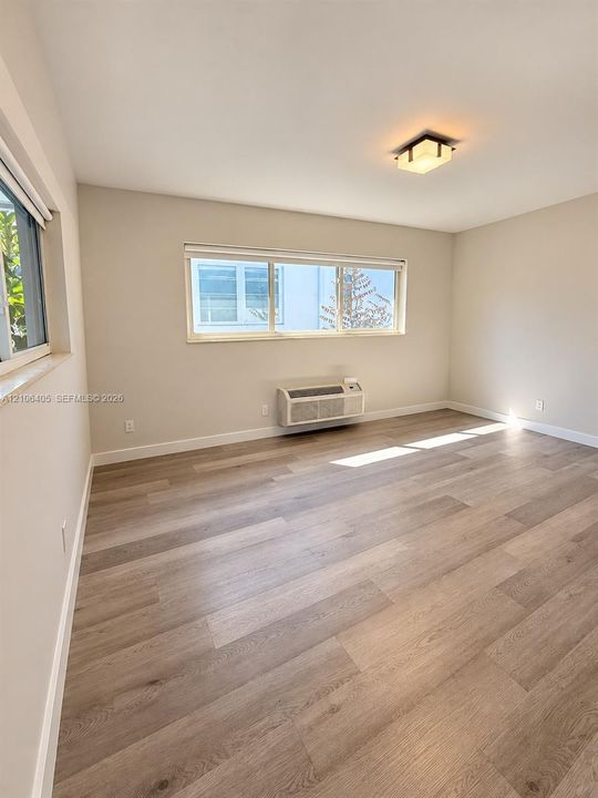 Neutral-toned interior featuring wood-finish flooring, dual horizontal sliding windows, a wall-mounted climate control unit, and a square flush-mount light fixture
