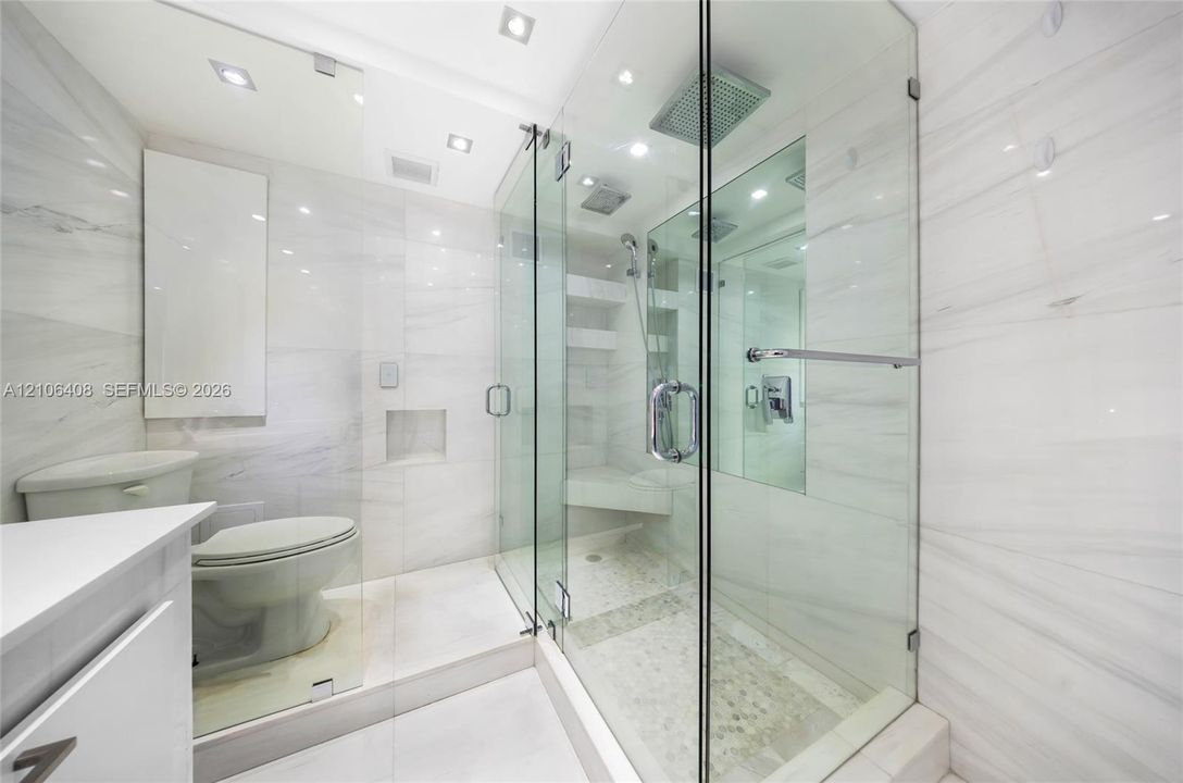 Frameless glass shower enclosure with rainfall showerhead, integrated tiled bench, pebble stone flooring, and floor-to-ceiling stone-tiled walls