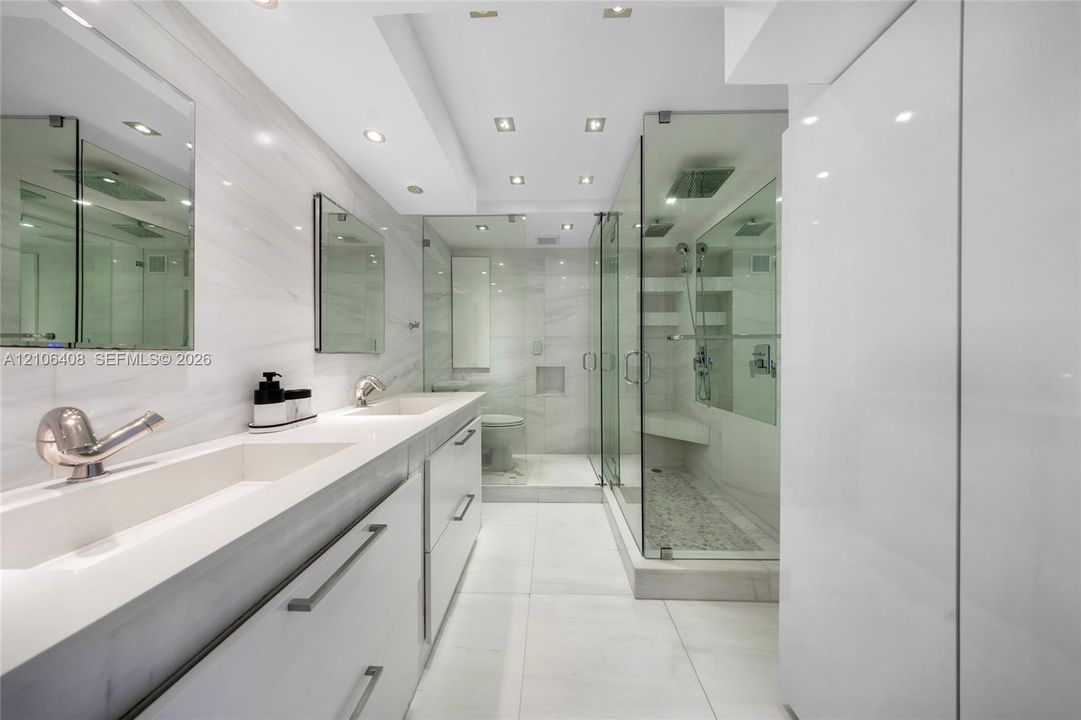 Dual-sink vanity with integrated basins and stone-finish countertops, glass-enclosed shower with built-in bench seating, large-format tile flooring, recessed lighting, and expansive wall-mounted mirrors