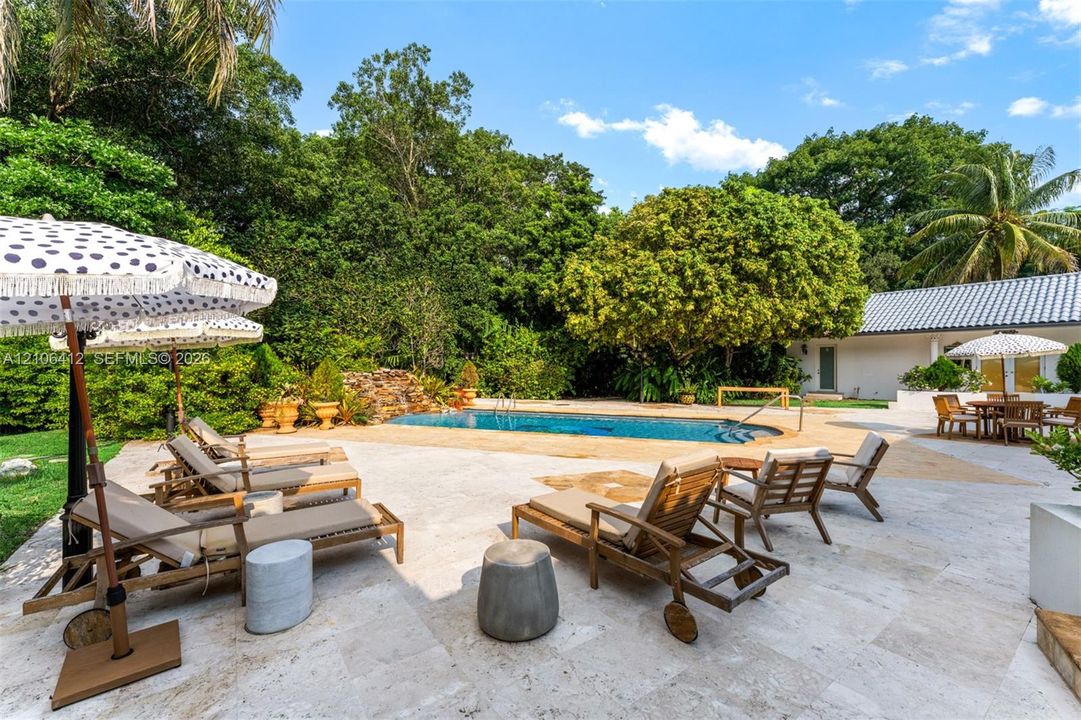 Expansive stone pool deck featuring a swimming pool with rock waterfall feature, mature perimeter landscaping, and tile roof structure