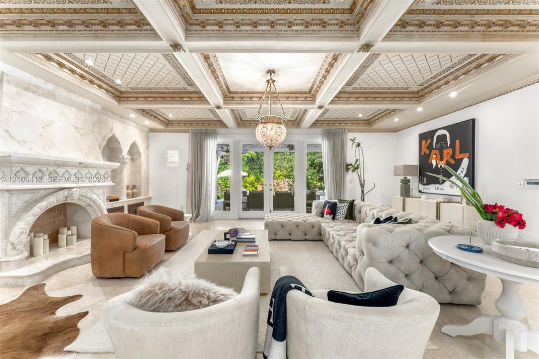 Ornate coffered ceiling with gold-leaf detailing, expansive stone fireplace with arched architectural trim, recessed lighting, and glass-paned double doors