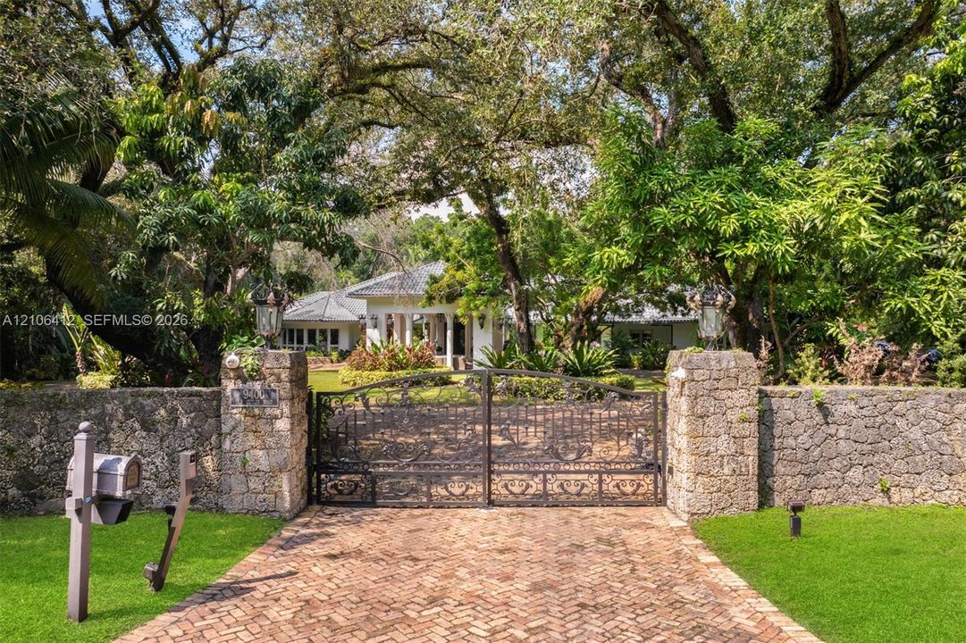 Gated estate entrance featuring a herringbone-patterned brick driveway, stone perimeter wall, ornamental metal gate, and mature canopy trees