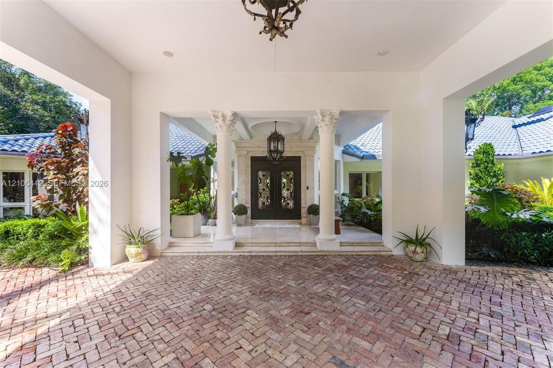 Covered motor court featuring herringbone-patterned brick paving, decorative classical columns, and dual entry doors with intricate metal detailing