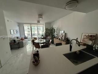 Open-concept interior featuring light-toned tile flooring, solid-surface kitchen countertops, an undermount sink with a gooseneck faucet, and floor-to-ceiling windows