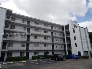 Multi-story apartment building exterior featuring private balconies, vertical architectural lines, concrete paved parking surface, and established perimeter shrubbery