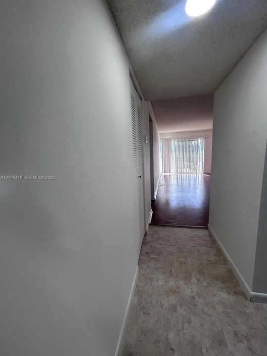 Hallway featuring neutral-toned tile flooring and a built-in louvered-door closet, leading to a space with dark-toned flooring and full-height vertical blinds